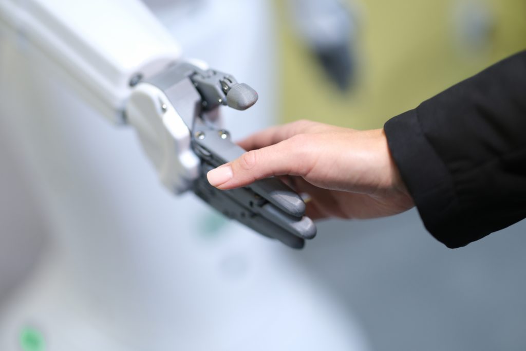 a person's hand, covered by a black long-sleeved shirt, is reaching out to shake hands with a silver robot with multiple parts. The image captures the meeting of two different worlds - the traditional human world and the emerging world of artificial intelligence.