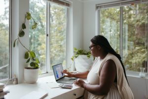 A dark skinned person with long braids interacts with their laptop on the left. Their office is bright with white walls, desk, supplies, and plant pot. Past the windows are relaxing greenery and trees.